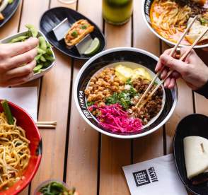 Spread of Asian-fusion foods on a wooden table with hands and chop sticks picking items off of plates and bowls.