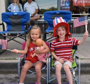 Two kids sitting in lawn chairs at a parade waving American flags