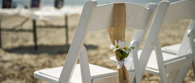 A white chair on a sandy beach is decorated with burlap and small white flowers, with a serene ocean backdrop.
