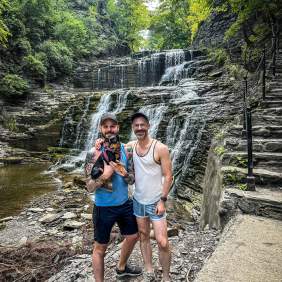 Two men stand in front of a waterfall, holding a small dog.