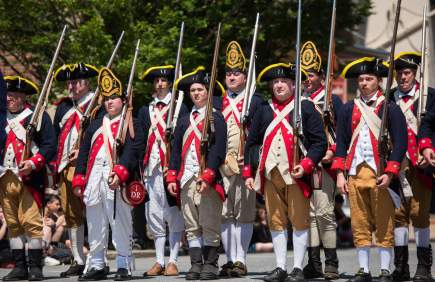 Historical reenactors in Colonial American military uniforms, holding muskets, standing in formation outdoors.