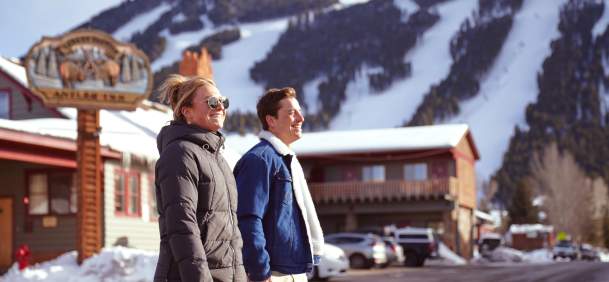 Two visitors walk through downtown Jackson, Wyoming, on a sunny winter day with snow-covered ski runs at Snow King Mountain rising behind them. The scene highlights Jackson’s walkable downtown, winter atmosphere and close connection to outdoor recreation in Teton County.