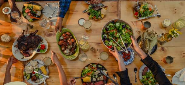 An overhead view of several people gathered around a wooden outdoor table, sharing a meal of grilled meats, corn, potatoes, and fresh salads in a warm evening setting.