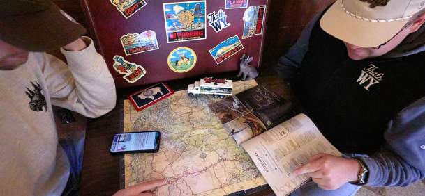 Two travelers plan a Wyoming road trip using a state map and Wyoming travel guide. Wyoming-themed stickers, a miniature truck and travel materials surround the map as one traveler points to a route, highlighting trip planning and exploration across the state.