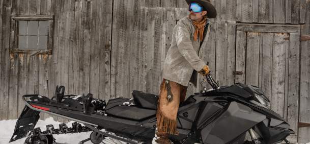 A cowboy dressed in winter Western attire stands on a snowmobile beside a weathered wooden barn near Jackson, Wyoming. Snow-covered chaps, a cowboy hat and ski goggles blend Wyoming’s Western heritage with modern winter recreation in Teton County.