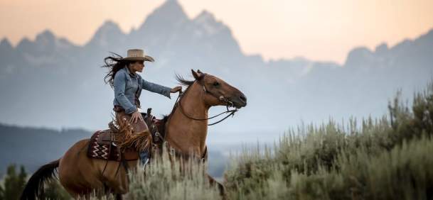 Woman riding her horse up a hill with the Grand Tetons in the background.