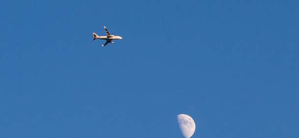 An airplane flying across a clear blue sky with the moon in view.