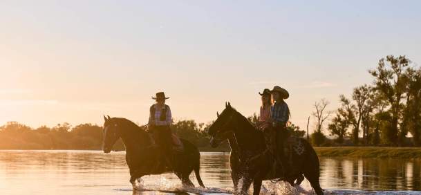 Three women on horse back wading through river with trees in the background.