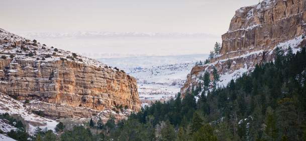 A vehicle travels along a snow-covered mountain road through a rugged canyon near Lander, Wyoming. Layered rock cliffs, evergreen trees and a broad winter landscape frame the roadway, highlighting a scenic winter drive through Fremont County.