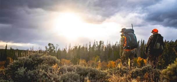 Two sportsmen with gear hiking throigh sage filled field at sunset.
