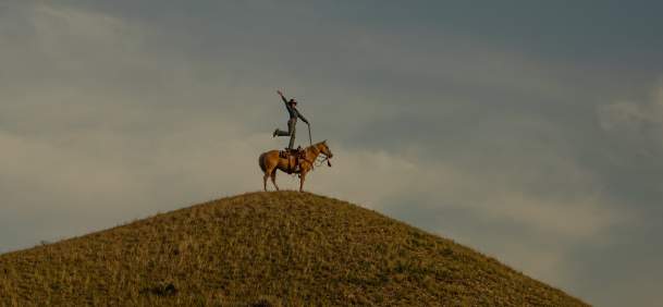 A rider stands with one arm raised on a horse at the top of a grassy hill, silhouetted against a wide sky with soft clouds.