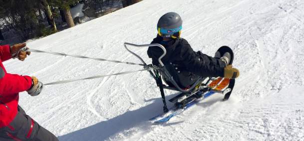 An adaptive skier uses a sit-ski while being assisted by an instructor on a snow-covered ski slope in Wyoming. The skier wears a helmet and winter gear as they navigate the mountain, highlighting accessible outdoor recreation and adaptive skiing opportunities.
