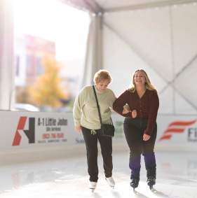Two women skate with their arms linked at Denison on Ice. The woman on the left is older with short hair, a cream colored sweatshirt, black pants, and a black crossbody purse. The woman on the right is younger with long blond hair, a brown cardigan, and black jeans.