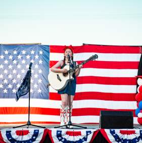 A young girl wearing white cowboy boots, a blue dress, and a red bow on her head plays guitar in front of an American flag backdrop at the 2026 Fourth of July Spectacular.