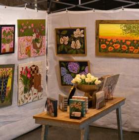 A vendor booth set up at the North Texas Art Festival. There are paintings of flowers and landscapes hung on white backdrops. A vase with white flowers and some smaller canvases are standing upright on a small table in the center of the booth.