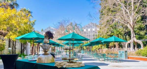 Event space of Mission Plaza and San Juan Villa at Gilroy Gardens shows outdoors event tables and umbrellas with decor and serving ware on a green linen lined table. The background is a blue sky with a large greenhouse and unique trees.