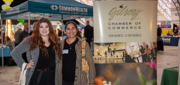 Under a massive event plaza tent, two smiling women stand next to a banner sign for the Gilroy Chamber of Commerce