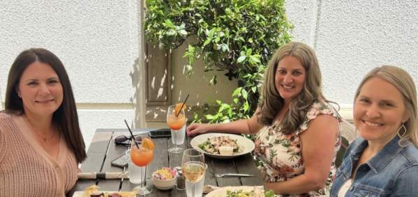 three ladies sit at a table outdoors with drinks and plates with food