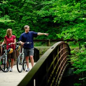 A person pointing while one stands with bicycles on a wooden bridge surrounded by lush green forest in the Gatlinburg scenic trail.