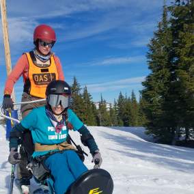 A skier uses adaptive ski equipment with a companion to ski the slopes. Snow, trees and blue sky in the backdrop.