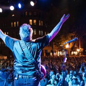guitar player on stage with hands up in front of huge crowd at night