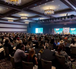 Interior of a ballroom in Fairmont Hotel with meeting attendees at round tables and a speaker on the stage at the front of the room