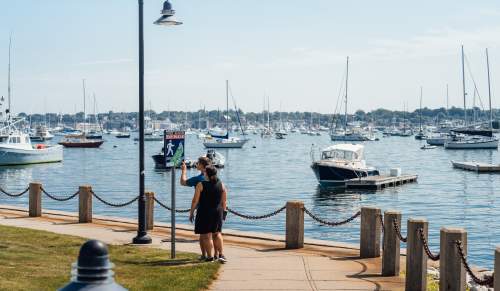 Waterfront walkway in Newport, RI with two people looking at a Fort Adams Bay Walk sign, with boats and sailboats docked in the harbor in the background.