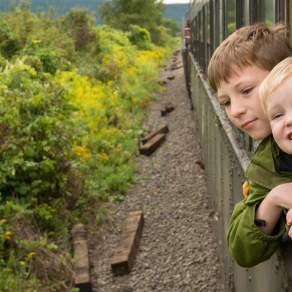 Two children leaning out of a train window with plant life to the left of the track.