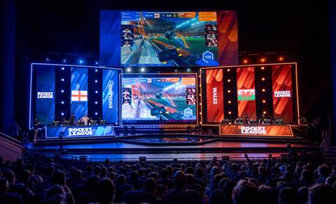 A crowd sit in darkness in a large venue in front of a stage lit in bright blue and red, ready to host an esports event