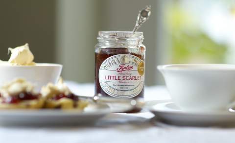 An open jar of Tiptree Littel Scarlet Jam sits on a table beside a bowl of cream, a cup of tea and some scones.