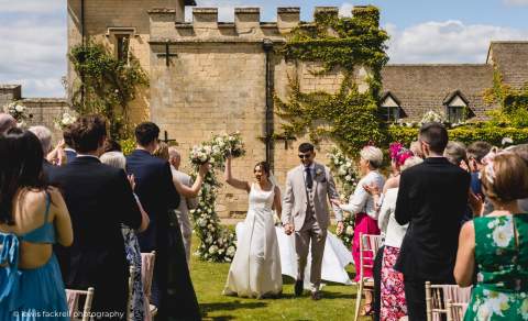 A couple smile at guests as they walk down the aisle at their outdoor wedding ceremony in the gardens of Ellenborough Park