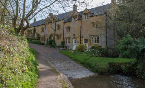 A row of traditional Cotswold stone cottages beside a narrow lane, with a shallow stream running alongside and trees and greenery in the foreground.