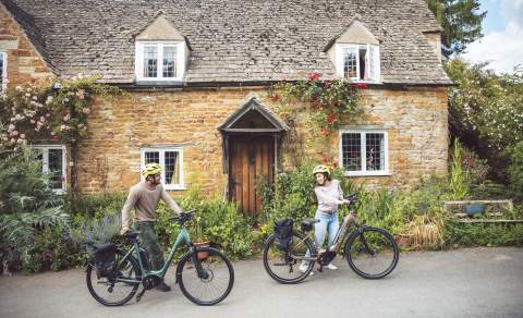 A couple push their bikes past a beautiful old cottage in a Cotswold village