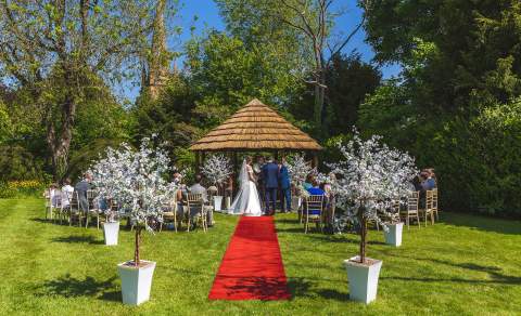 A couple getting married infront of a wooden gazeebo, with friends and family seated on chairs, a red carpet leading away to decorative blossom trees