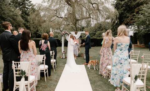 A bride and groom exchange vows at an outdoor ceremony, watched by their wedding guests and a Cavalier King Charles spaniel
