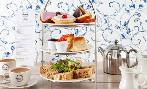 A three-tier cake stand filled with finger sandwiches, scones and delicate cakes sits on a table with a teapot and teacups at Huffkins Café & Bakery in the Cotswolds