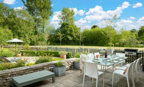 A sunny outdoor patio set with a glass-topped dining table surrounded by white chairs, prepared for a meal with plates, glasses, and napkins. The patio overlooks a neatly kept garden with potted plants, a low stone wall, and a picket fence. Beyond the fence, there is a grassy field bordered by tall, leafy trees under a bright blue sky with scattered clouds.