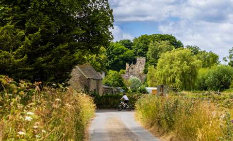 A cyclist viewed down a deserted road with a church and houses in the distance