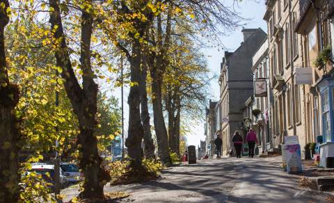 Dappled sunlight casts shadows on the pavement as people walk along High Street, Witney in the Cotswolds