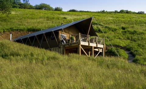 A Safari tent with a wooden deck sits on a lush green hillside under a clear blue sky. The scene conveys tranquillity and an outdoor adventure vibe.