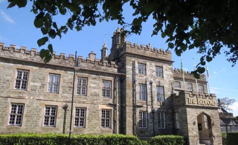 Historic stone building with crenellated roofline and tall windows, labeled "The Bedford." Framed by tree branches, evokes a sense of timeless elegance.