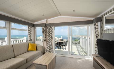 Bright living room with a beige sofa and yellow cushion, facing sliding glass doors that open to a balcony with a sea view. Airy and serene atmosphere.