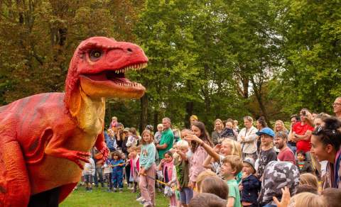 A vibrant red and orange dinosaur costume entertains an excited group of children and adults in a lush park, creating a fun and playful atmosphere.