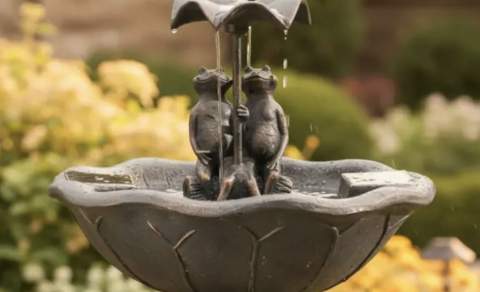 A stone fountain with two frog sculptures holding an umbrella, surrounded by lush garden foliage. It's a serene and whimsical garden scene.