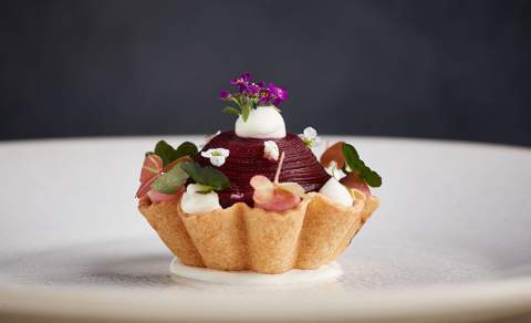 Elegant tartlet on a white plate featuring a beet sphere, dollops of white cream, edible flowers, and microgreens, set against a dark background.