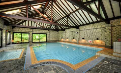 Indoor pool with rustic wooden beams and brick walls, sunlit through large windows. The pool is calm, with seating nearby, creating a serene atmosphere.