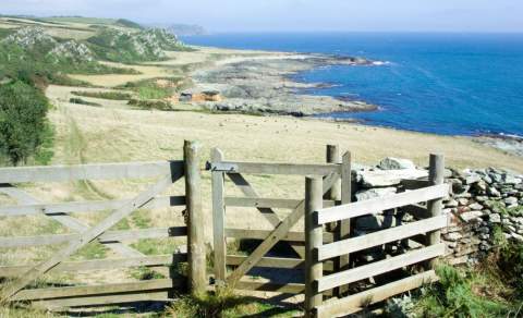 A wooden gate opens to a coastal path with distant green hills and a vast, calm blue sea. The scene conveys a serene, inviting atmosphere.§