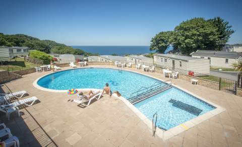 Outdoor pool with loungers, people sunbathing, and a colourful inflatable under a clear blue sky. Mobile homes and sea view in the background.