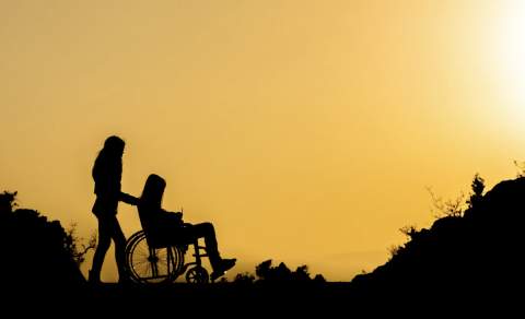 Silhouetted scene of a person pushing another in a wheelchair at sunset, set against an orange sky. Evokes themes of care and companionship.