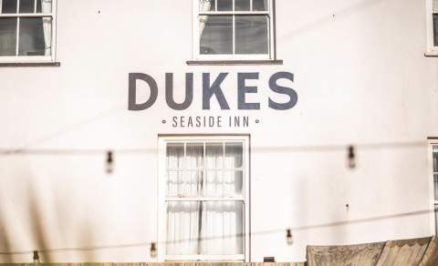 Facade of Dukes Seaside Inn with bold signage. The building features white walls and a curtained window. A relaxed, coastal ambiance is conveyed.